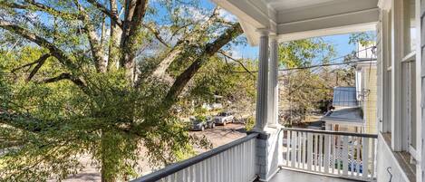A covered porch with a railing, overlooking a street with parked cars and a large tree.