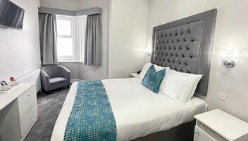 Hotelkamer met een groot bed, een grijs gestoffeerd hoofdeinde, een grijze fauteuil, een nachtkastje en een raam met gordijnen.