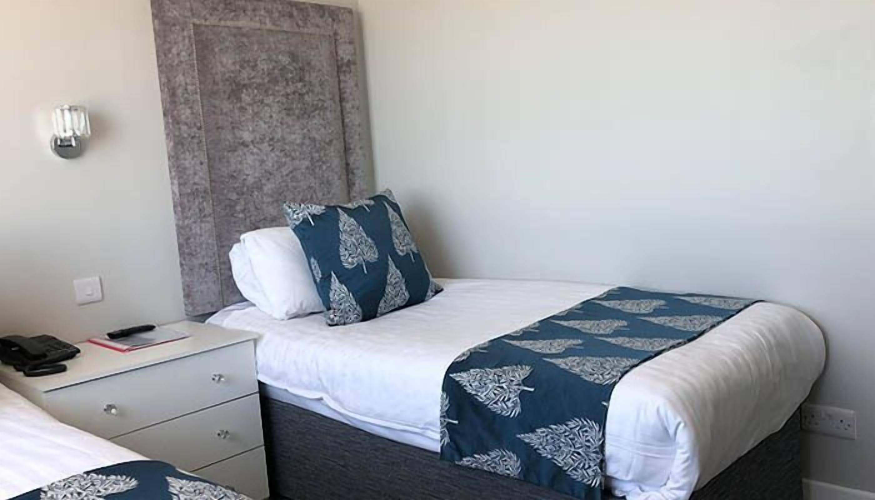 A neatly arranged hotel room with a bed, a bedside table with a phone and remote, and a wall-mounted light.