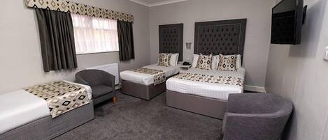A hotel room with two single beds, a double bed, a grey armchair, a small table, and a television mounted on the wall.