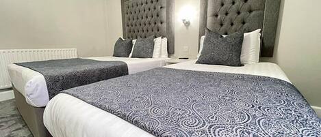 Habitación de hotel con dos camas, cada una con cabecero gris y ropa de cama blanca.