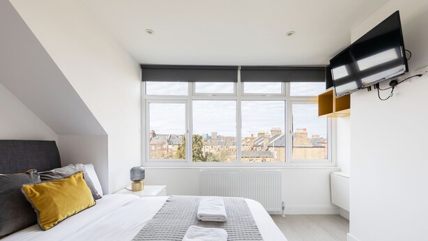 Un dormitorio moderno con un ventanal grande, un televisor de pantalla plana, una cama con ropa de cama blanca y gris, y vistas a la ciudad.