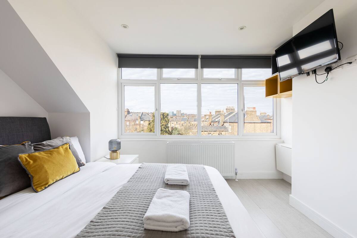 Un dormitorio moderno con un ventanal grande, un televisor de pantalla plana, una cama con ropa de cama blanca y gris, y vistas a la ciudad.