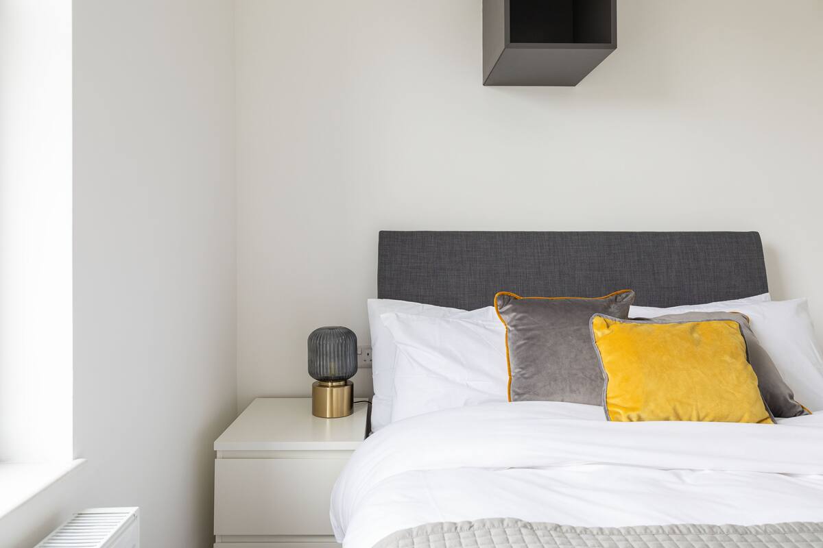 Una cama bien hecha con ropa de cama blanca y gris, una lámpara de noche y un pequeño estante sobre la mesita.