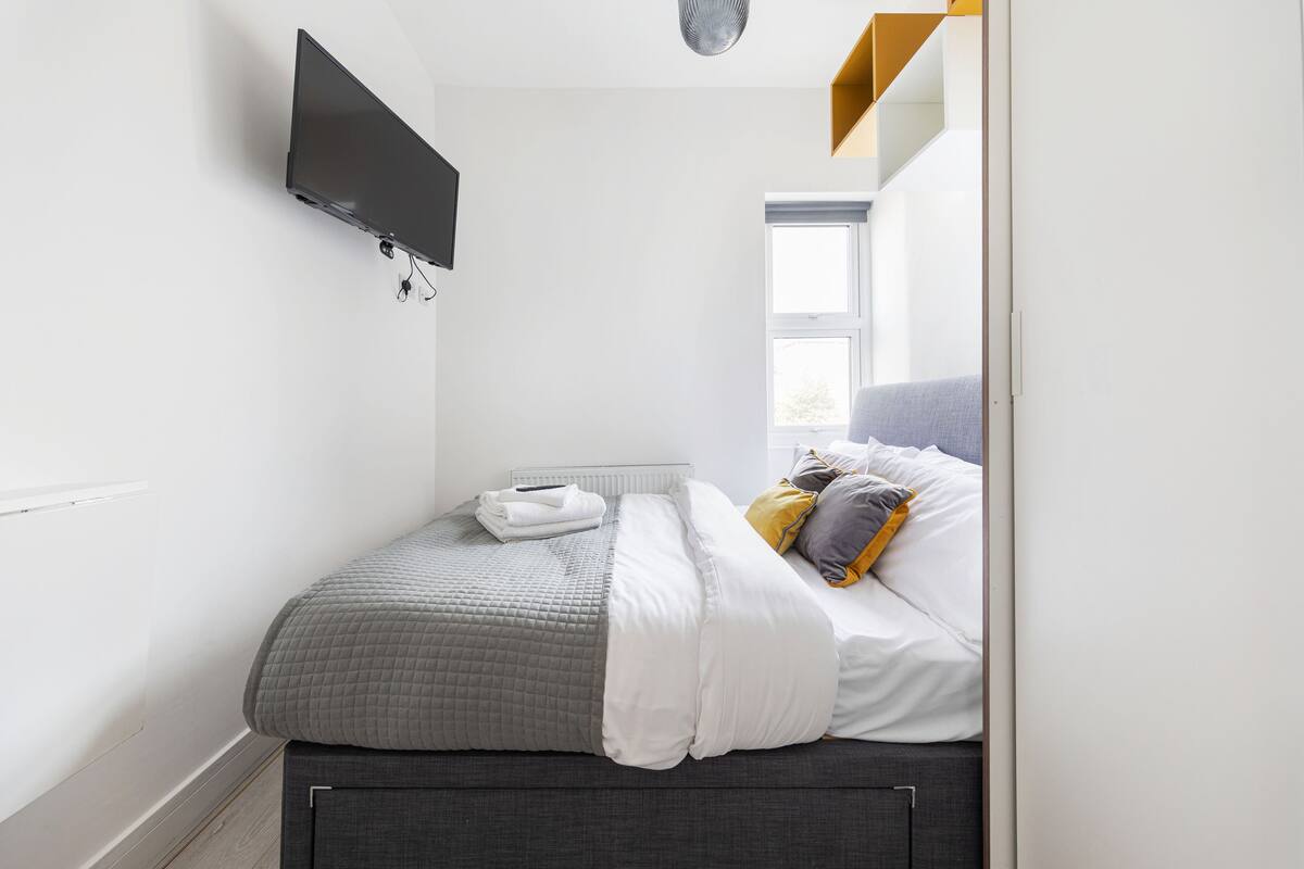 Un dormitorio pequeño y moderno con cama, televisor de pantalla plana montado en la pared y ventana que proporciona luz natural.