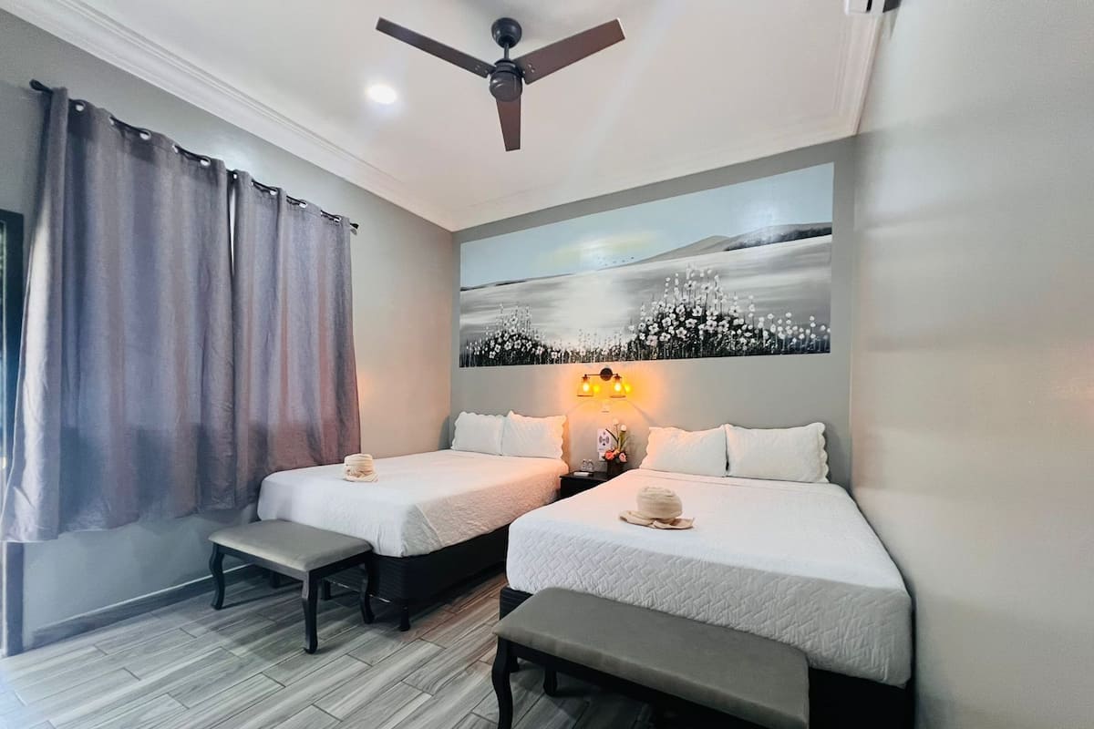 A hotel room with two beds, a ceiling fan, a painting on the wall, and a window with curtains.