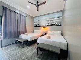 A hotel room with two beds, a ceiling fan, a painting on the wall, and a window with curtains.