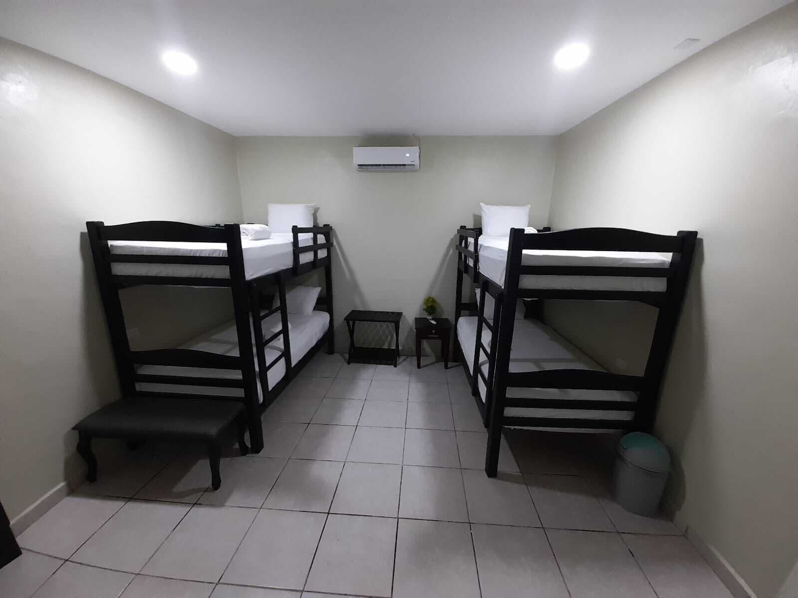 A dormitory room with two bunk beds, a small table, a trash bin, and an air conditioning unit on the ceiling.