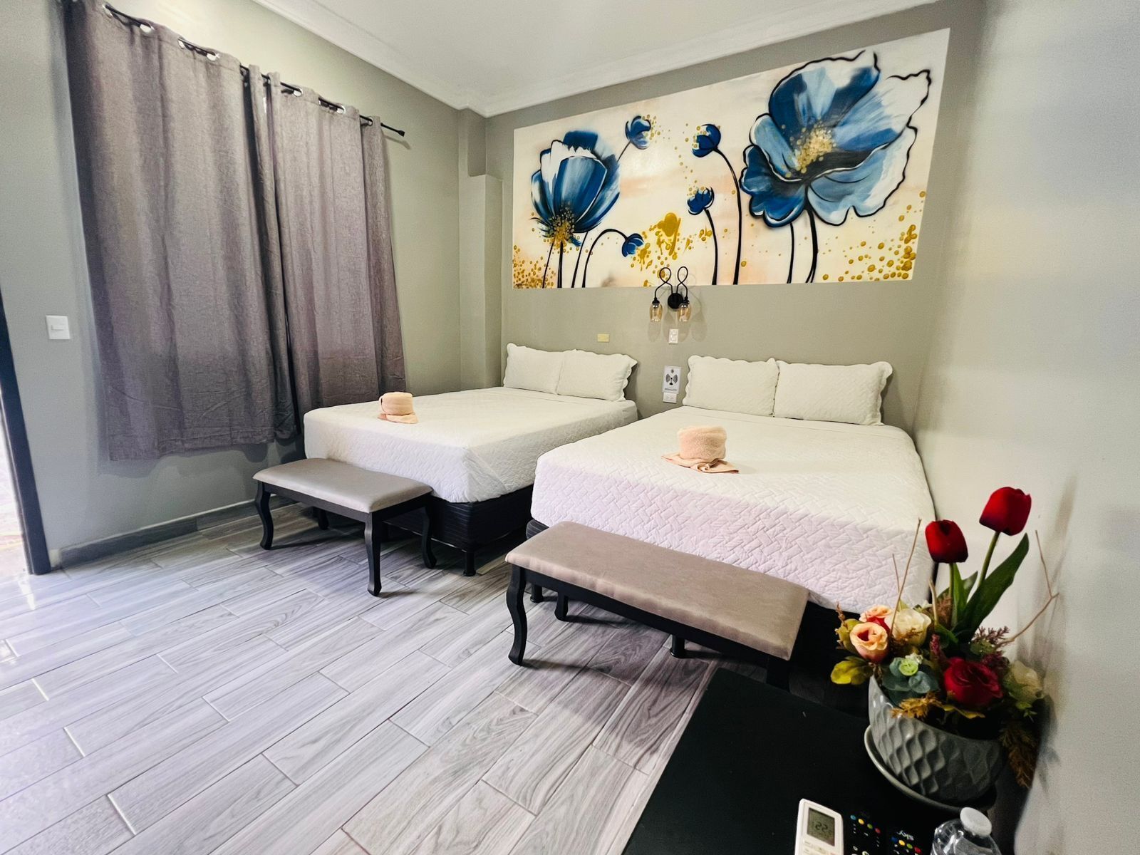 A hotel room with two beds, a bench, a wall-mounted clock, and a floral arrangement.