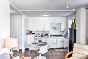 Une cuisine moderne avec des armoires blanches, un four à micro-ondes, une cuisinière et un réfrigérateur. On y trouve un coin repas avec une table et des chaises, ainsi qu’un élément décoratif mural au-dessus de l’évier.