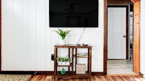 Una sala de estar con televisor montado, una consola de madera con plantas y una puerta que da a otra habitación.