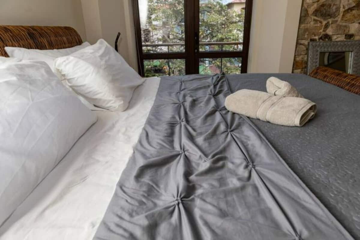 Una cama bien hecha con edredón gris y blanco, una toalla doblada y vistas al exterior a través de una ventana.
