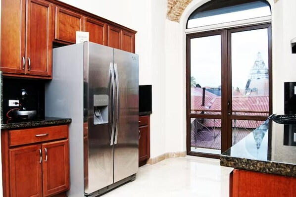 A kitchen with wooden cabinets, a stainless steel refrigerator, and a large window with a view of a building. - Los Diamantes (Panama City)