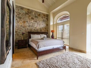 Un dormitorio con pared de piedra, una cama grande, una mesita de noche, una lámpara y vistas a través de un ventanal arqueado.
