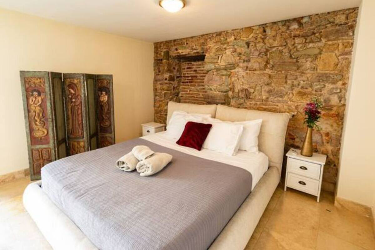 Un dormitorio con pared de piedra, cama con manta gris, almohadas blancas y una almohada roja. Hay una mesita de noche con un jarrón de flores y una toalla doblada sobre la cama.