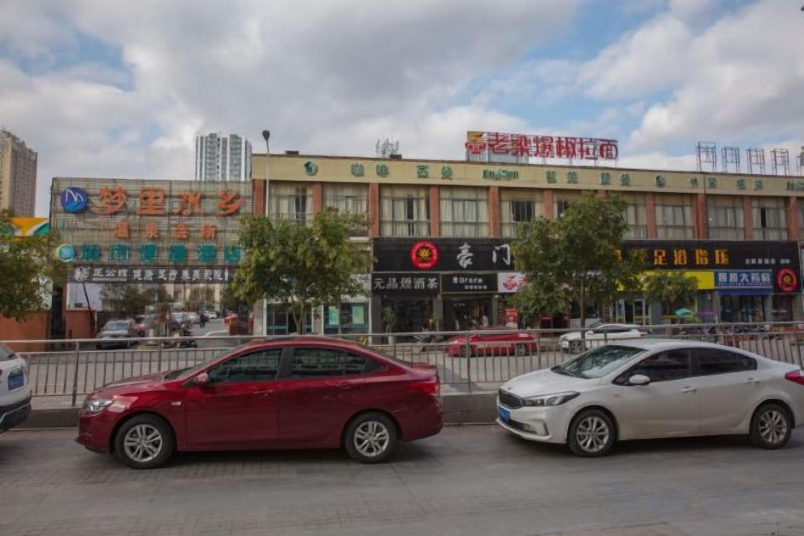 A street view with cars parked along the curb, buildings with commercial signage, and a clear sky.