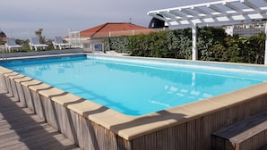 A rectangular swimming pool with a wooden deck surrounding it, a white pergola structure, and a red-roofed building in the background.