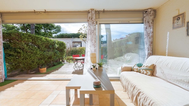 A room with a sofa, a table, and a view of a garden and buildings outside. - Appartement with terrace, wifi, parking (Agde)