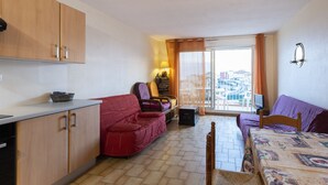 A compact living space with a kitchenette, a red sofa, a wooden dining table, and a balcony with a view. - Studio 4 ppl, sea view balcony, pool, parking (Agde)