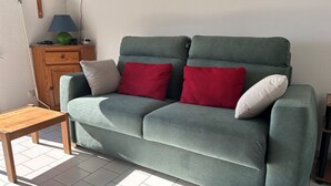 A green sofa with two red and two white cushions, a wooden side table, and a wooden cabinet with a lamp. - Studio 4 ppl, sea view terrace, private parking (Agde)