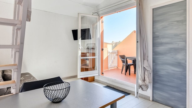 A room with a dining table, a black wire basket, a white wall, a sliding door to an outdoor area with a table and chairs, and a small shelf unit.