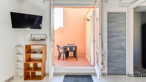 A room with a TV mounted on the wall, a wooden shelf unit, a table with two chairs, and a door leading to another room.