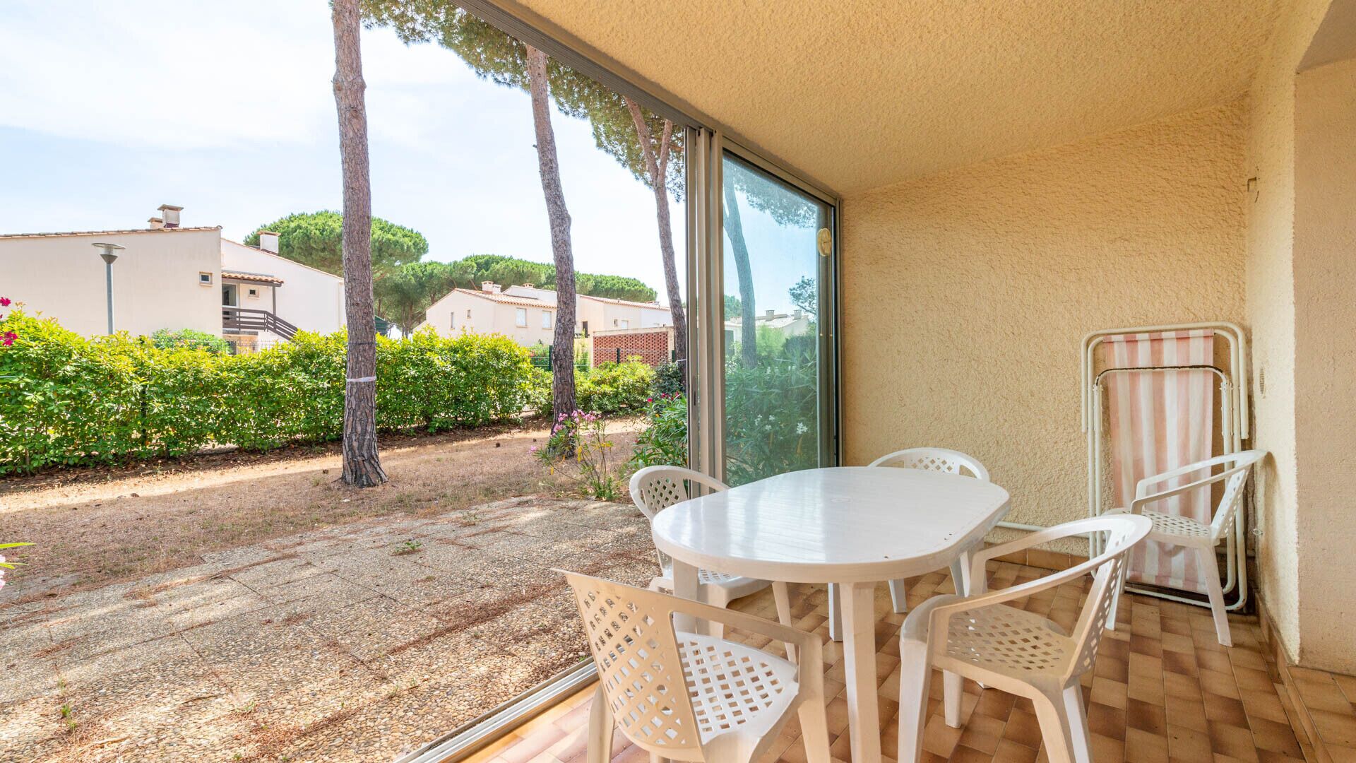 Une terrasse avec une table ronde et des chaises, une porte vitrée et une vue sur un jardin avec des arbres et un bâtiment en arrière-plan.