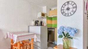 Uma cozinha compacta com micro-ondas, cafeteira e máquina de lavar loiça. Existe uma área de refeições com mesa e cadeiras, e um relógio de parede grande.