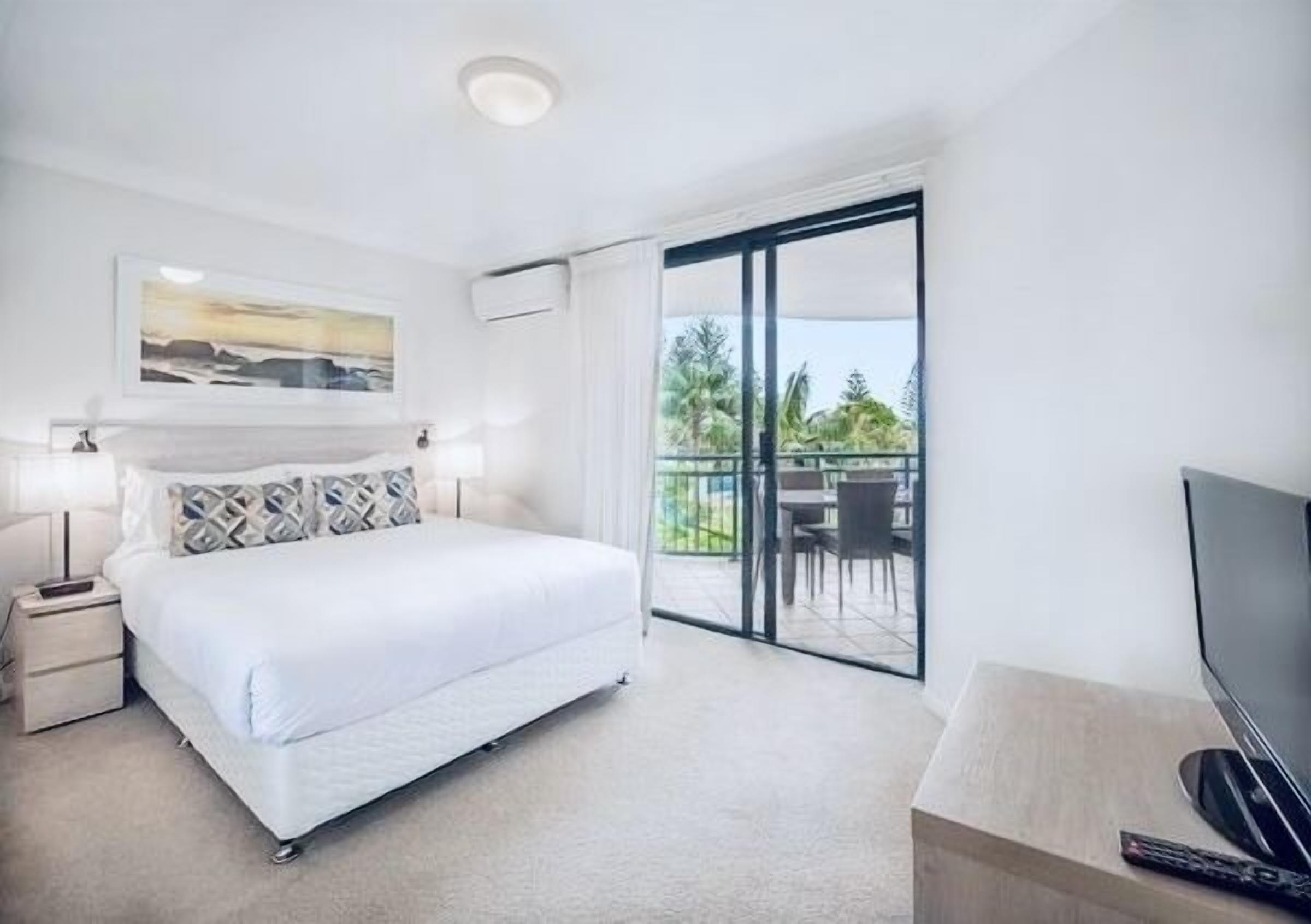 A hotel room with a large bed, a TV, a balcony with a table and chairs, and a painting on the wall.