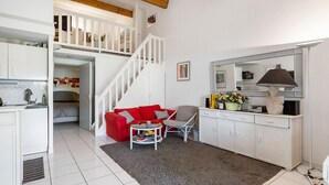 Een ruimtelijk ingerichte leefruimte met kitchenette, een rode bank, een kleine tafel en een trap naar een hoger niveau.