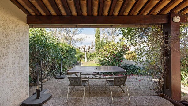 Een terras buiten, met een tafel en stoelen, omgeven door groen en uitzicht op een tuin.
