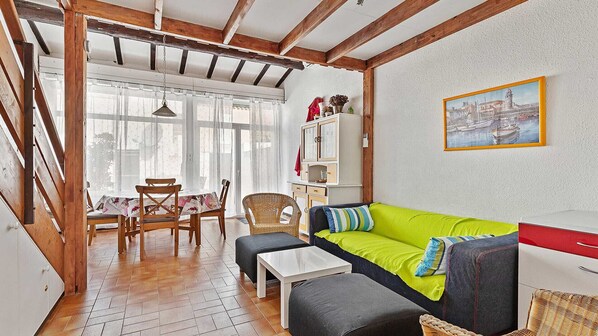 Um quarto com teto de madeira, um sofá com almofadas, uma mesa de jantar com cadeiras e uma janela com cortinas leves.