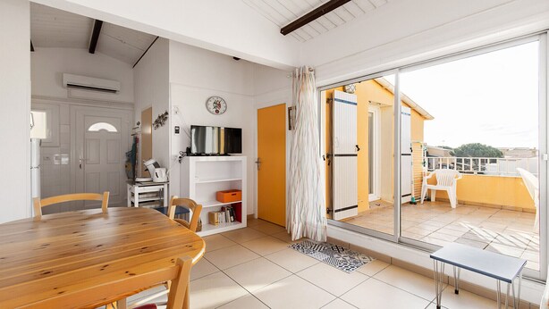 Una stanza con un tavolo da pranzo in legno, una TV su uno scaffale bianco, una porta-finestra in vetro scorrevole su un balcone e un condizionatore d'aria bianco.