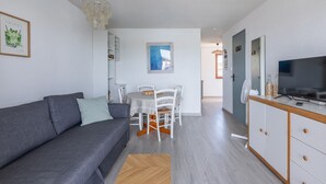Un salon moderne avec un canapé gris, une table à manger avec des chaises blanches, un téléviseur posé sur un meuble en bois et un petit tapis.