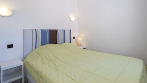 Una cama individual con colcha verde, una mesita de noche blanca, una lámpara y un aplique de pared.