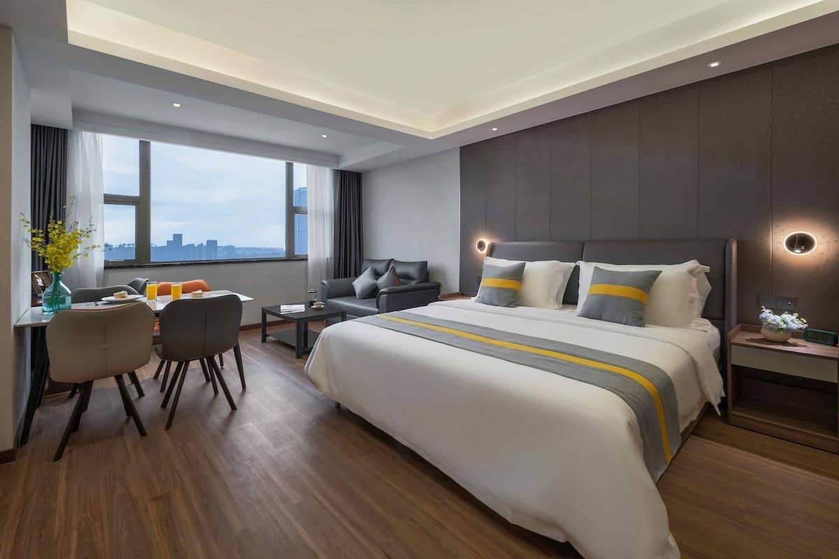 A modern hotel room with a large bed, a desk with chairs, a small table, and a nightstand with a lamp.