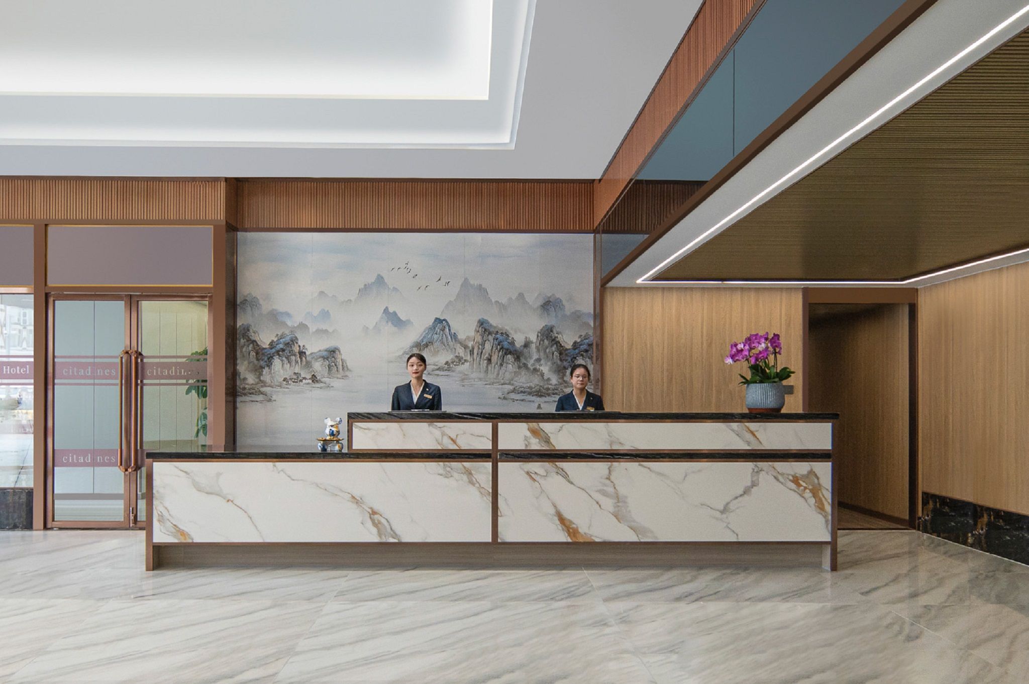Zona de recepción de un hotel con mostrador de mármol, dos recepcionistas, un cuadro grande de montañas y una planta en maceta con flores moradas.