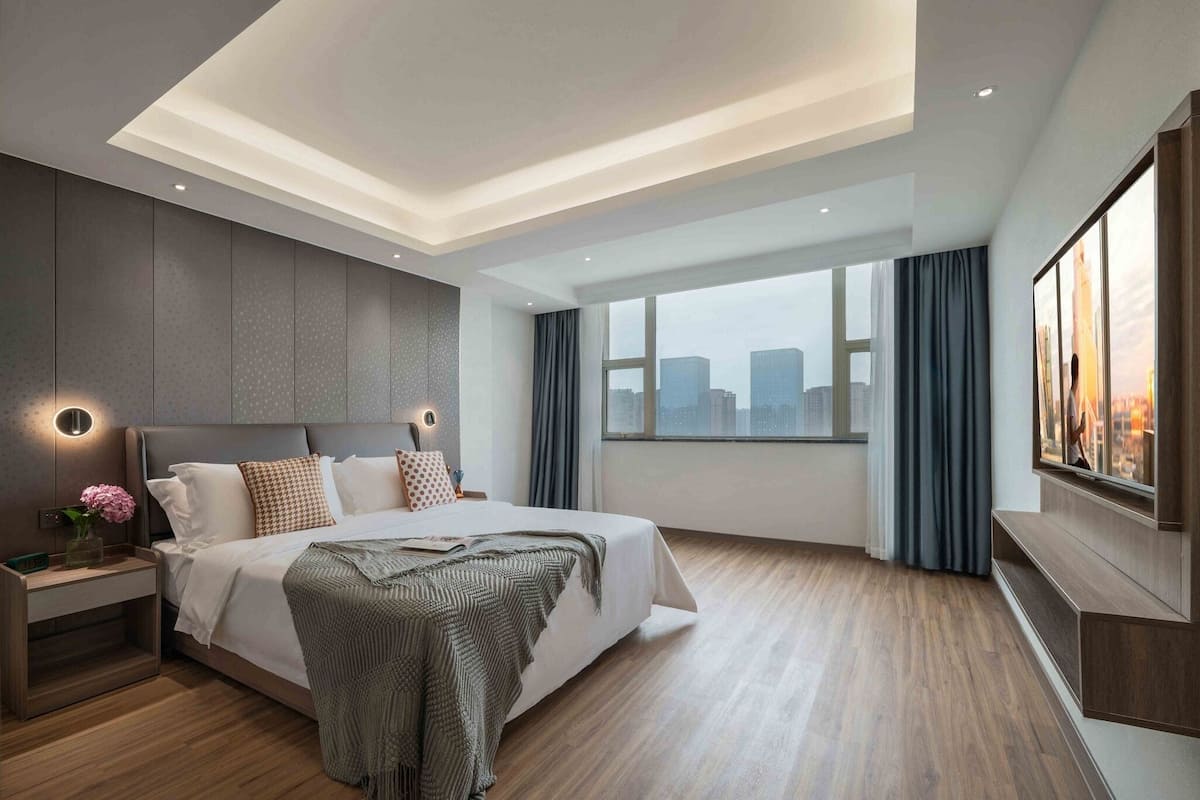 A modern hotel room with a large bed, a nightstand, a TV, and a city view.