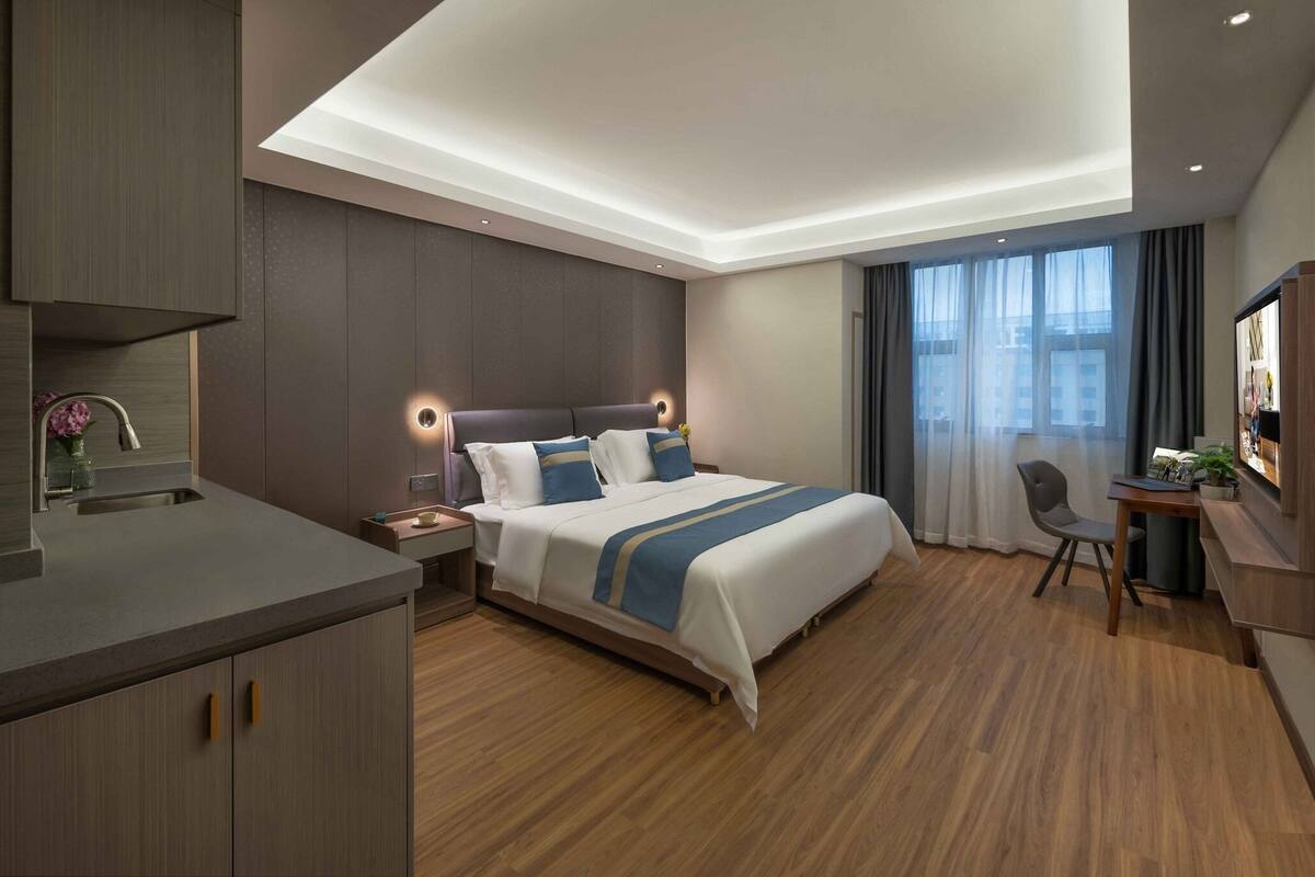 A modern hotel room with a large bed, a desk, a chair, and a small kitchenette.