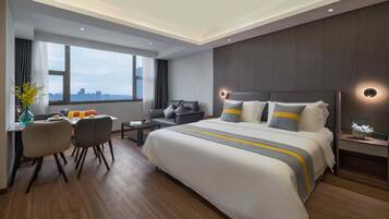 Una habitación de hotel moderna con una cama grande, un escritorio con sillas, una mesita pequeña y una mesita de noche con lámpara.
