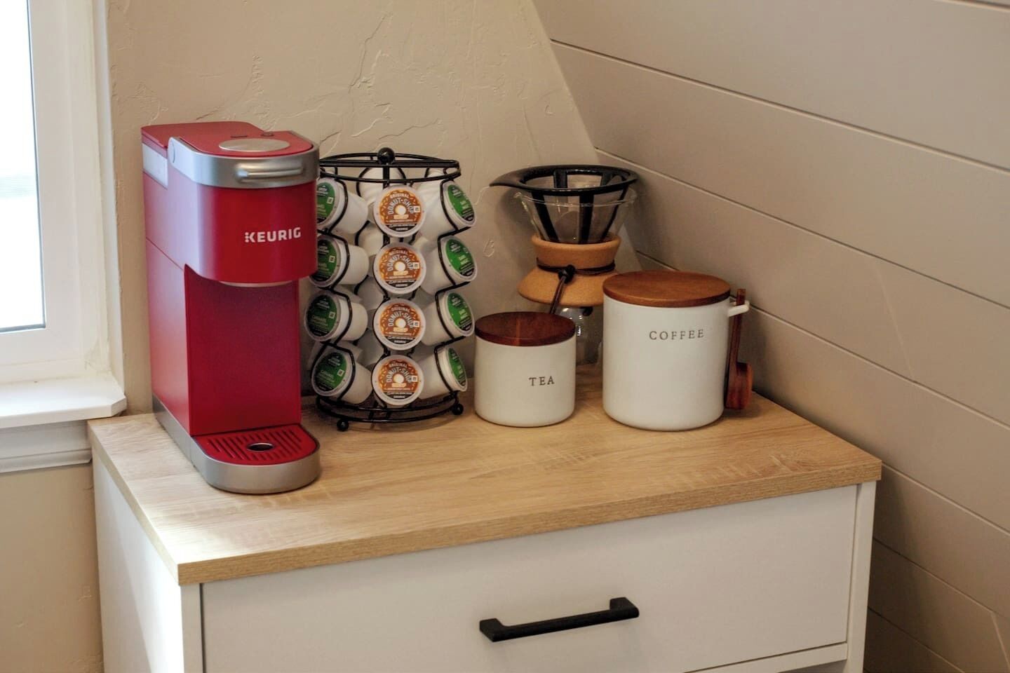 Una kitchenette con una cafetera Keurig roja, un molinillo de café y dos recipientes etiquetados como 'té' y 'café' sobre una encimera de madera.