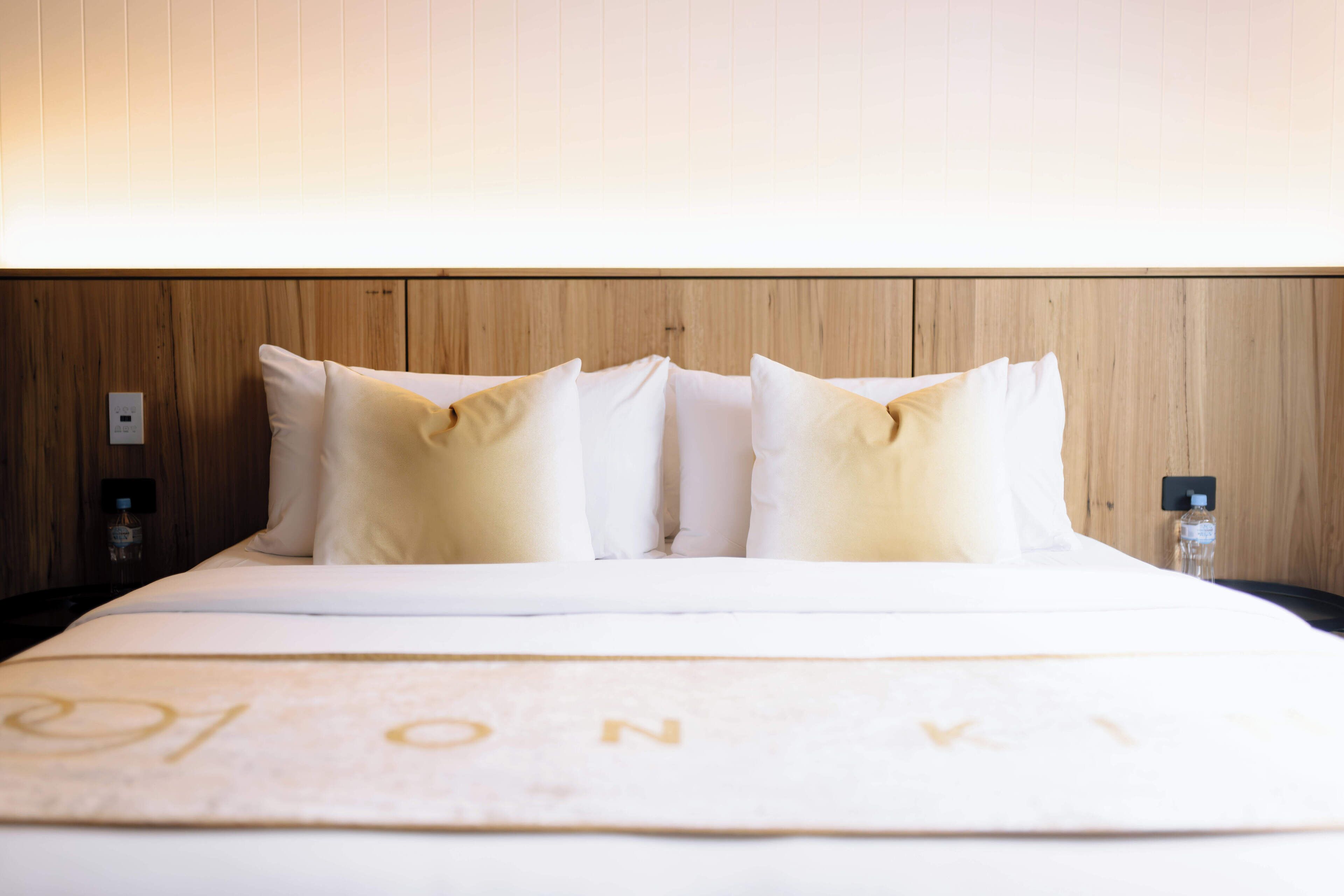 Un lit bien fait, avec une literie blanche et deux oreillers, une tête de lit en bois et un luminaire fixé au mur.