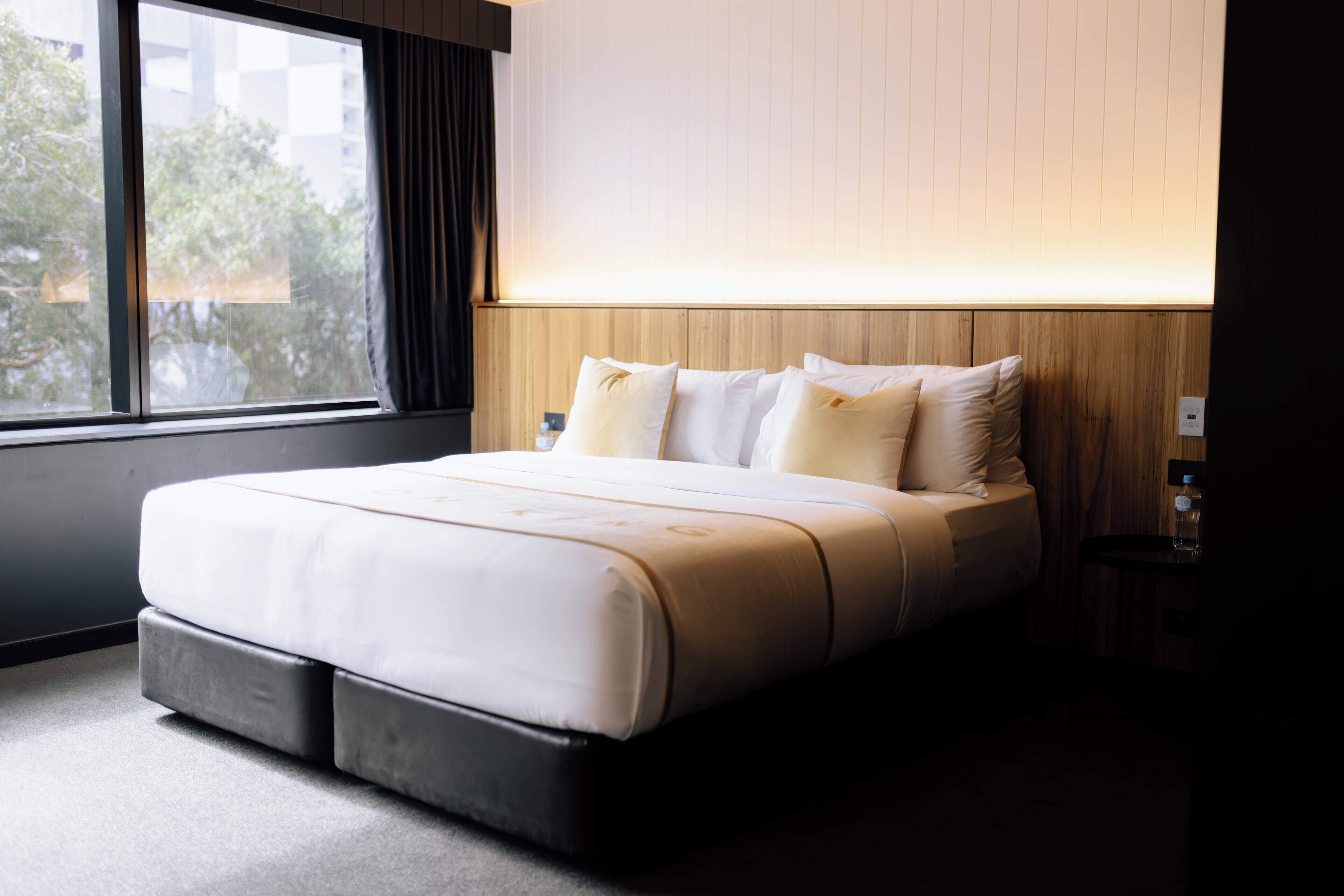 A modern hotel room with a large bed, a wooden headboard, a nightstand, and a window with curtains.
