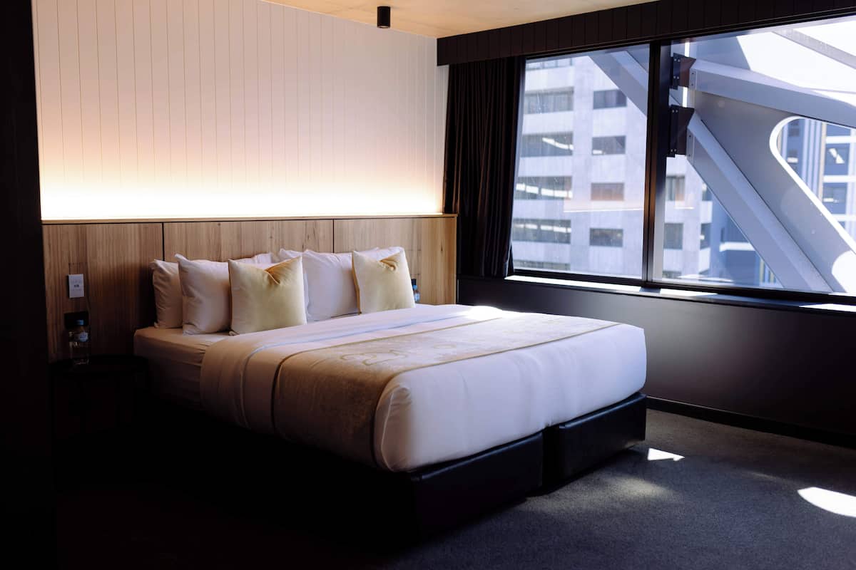 A modern hotel room with a large bed, a nightstand, and a window with a view of a building and a staircase.