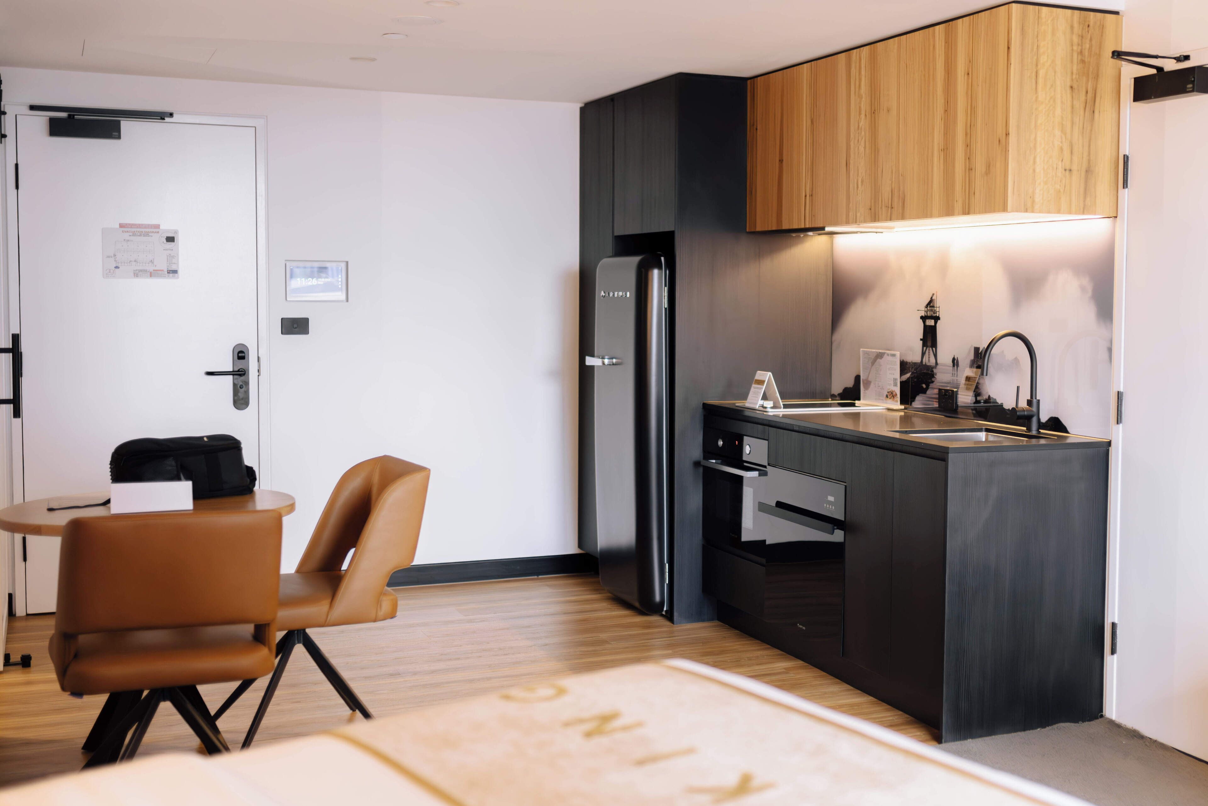 Cocina compacta con un refrigerador negro moderno, armarios de madera y un pequeño comedor con una silla marrón y una mesa.