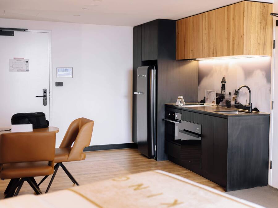 Cocina compacta con un refrigerador negro moderno, armarios de madera y un pequeño comedor con una silla marrón y una mesa.