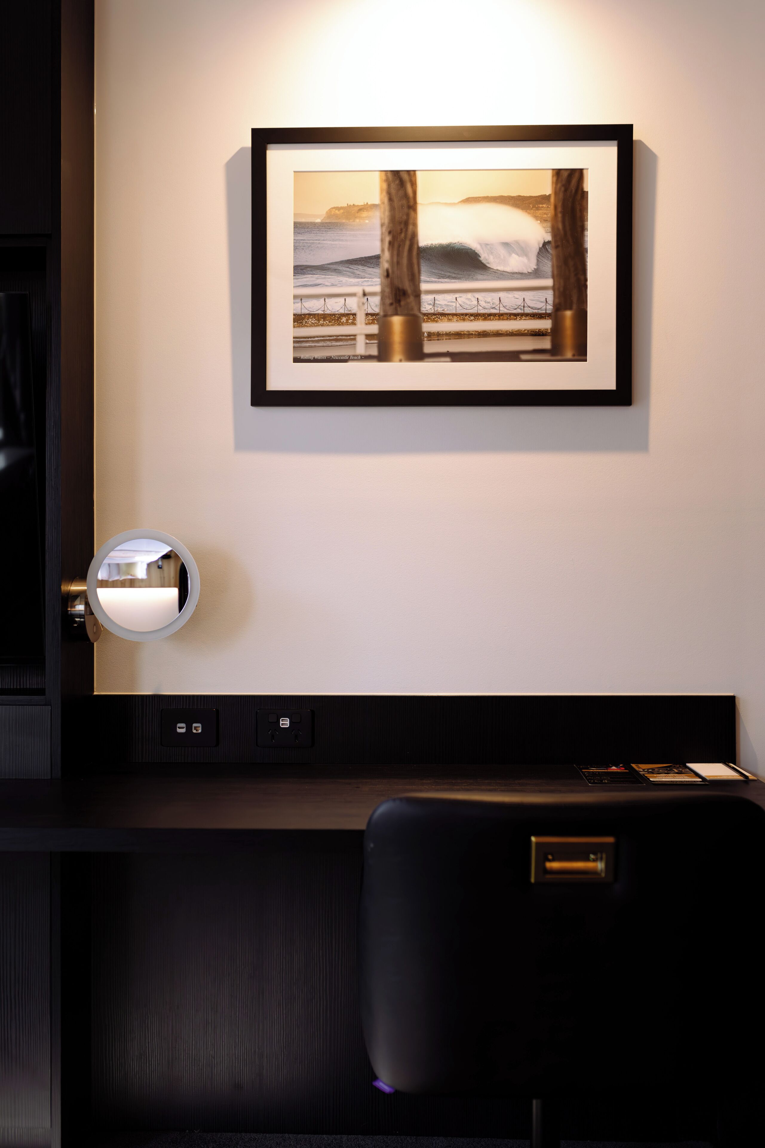 A framed picture of a coastal scene on a wall, a round mirror, and a dark-colored headboard with a control panel.