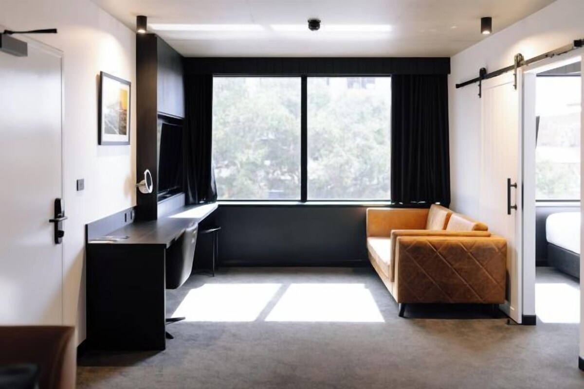 A modern hotel room with a desk, a sofa, and a large window offering a view of trees.