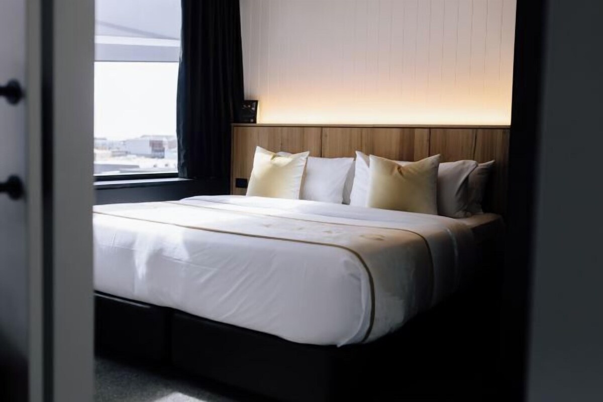 A neatly made bed with a wooden headboard, white bedding, and a window with a view of buildings.