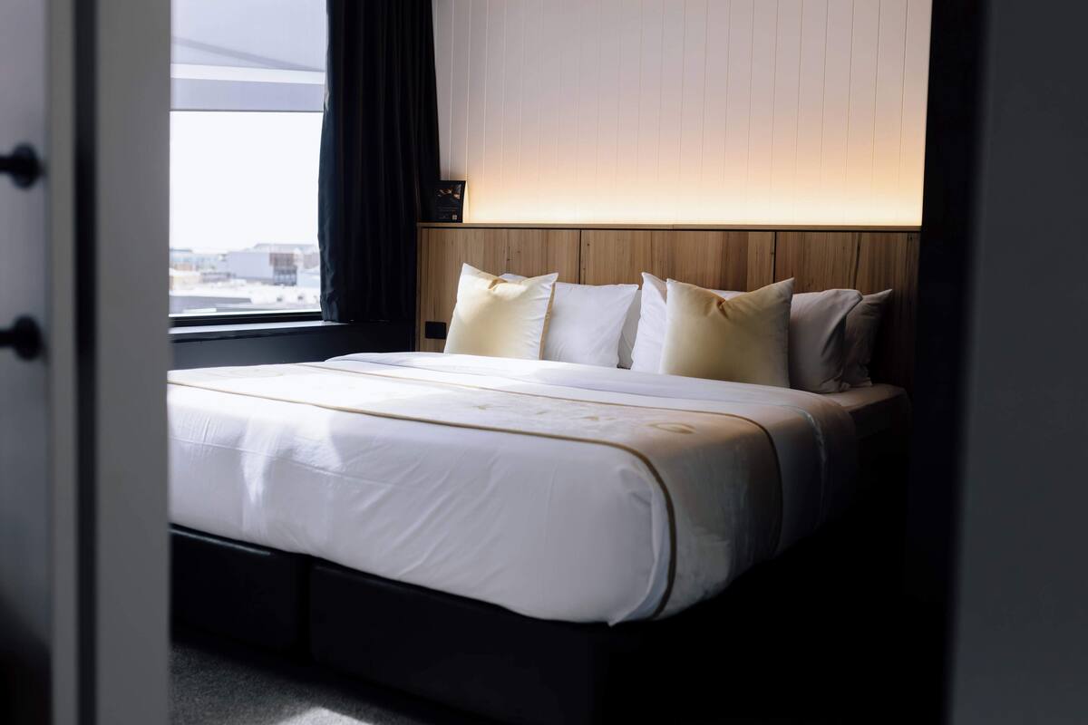 A neatly made bed with white linens and multiple pillows, a wooden headboard, and a window with a view of buildings.